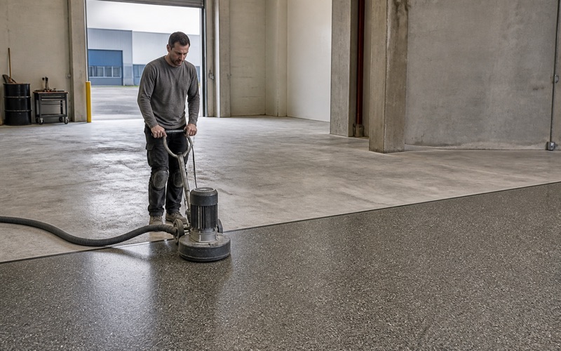 A worker forms a coved resin base at the foot of a stainless kitchen line, the seamless gray floor falling to a flush stainless drain.